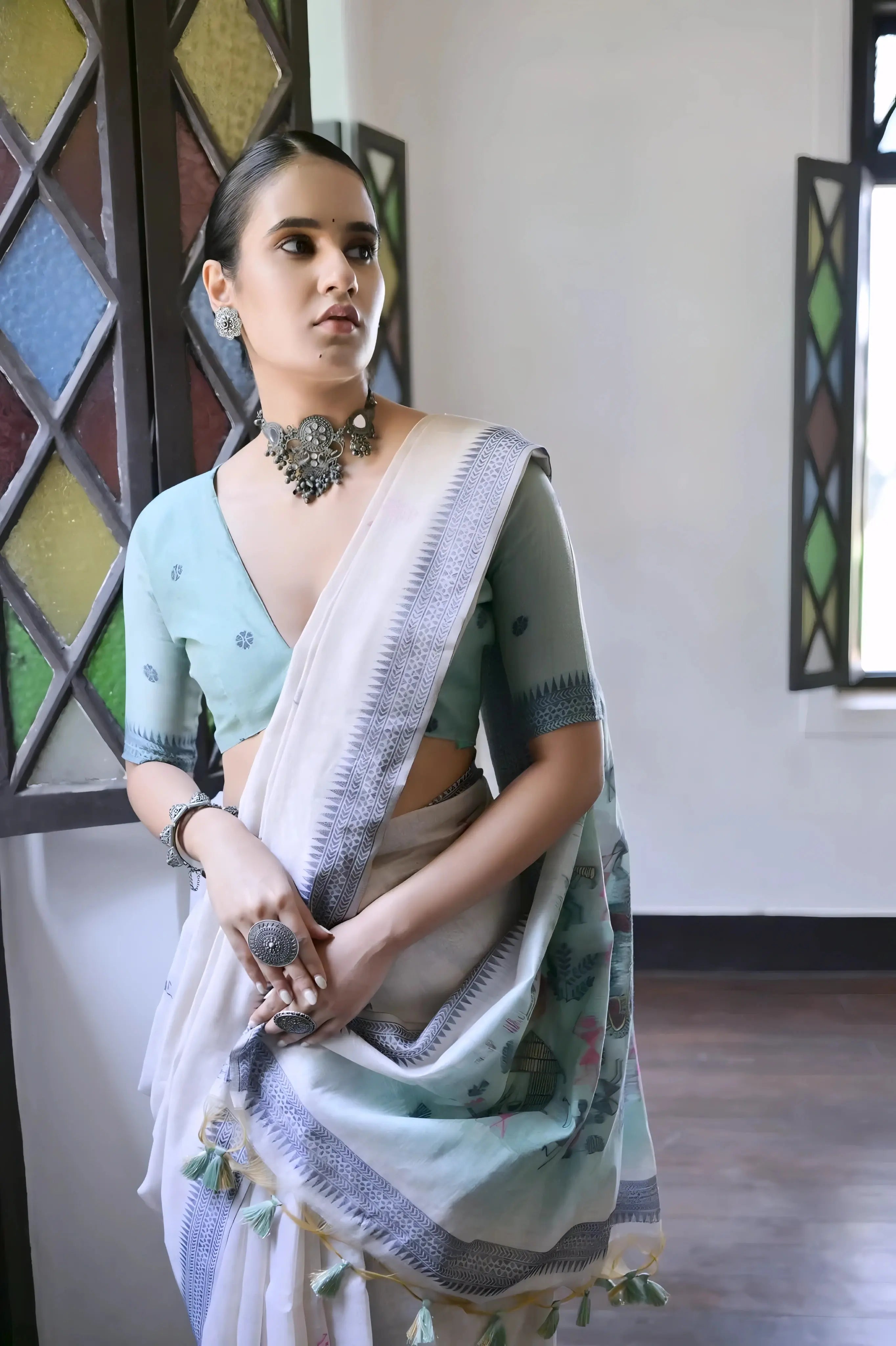 A woman models a graceful Off White Soft Muga Cotton Silk Saree. The pallu features a wide sea green panel decorated with traditional Warli-style figurative motifs in pink and red. She wears a light blue blouse and a dark oxidized choker necklace, posing indoors.